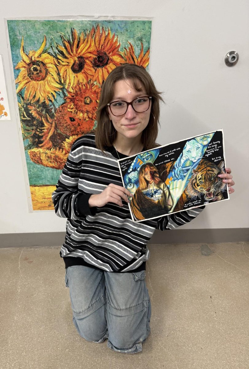 Senior Zoey Hebert posing with her artwork. “This piece is about how I use my voice to people in my everyday life,” Hebert said. “The blue strips are supposed to represent the ‘at-home’ aspect and the development. The more warm colored sections are supposed to represent how I am at school. That's why I'm wearing the hoodie.” Photo courtesy of Natalie Jones